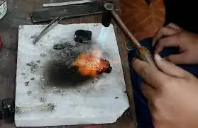 A person is using a torch to melt and shape an orange-colored metal piece on a white workbench.