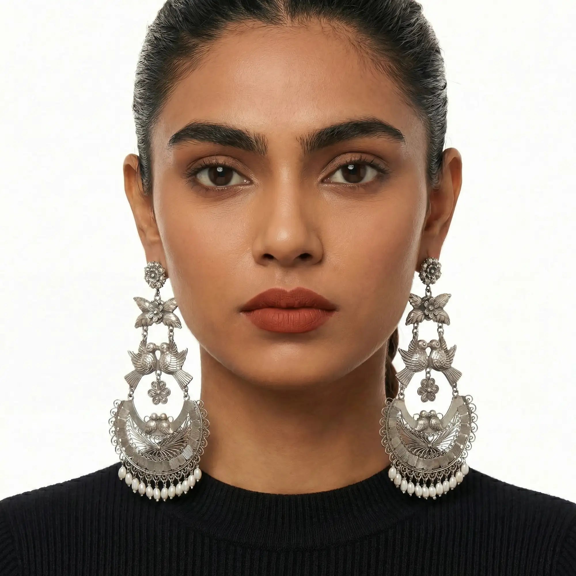 Silver, intricately designed chandelier earrings featuring birds and floral motifs with dangling white beads.