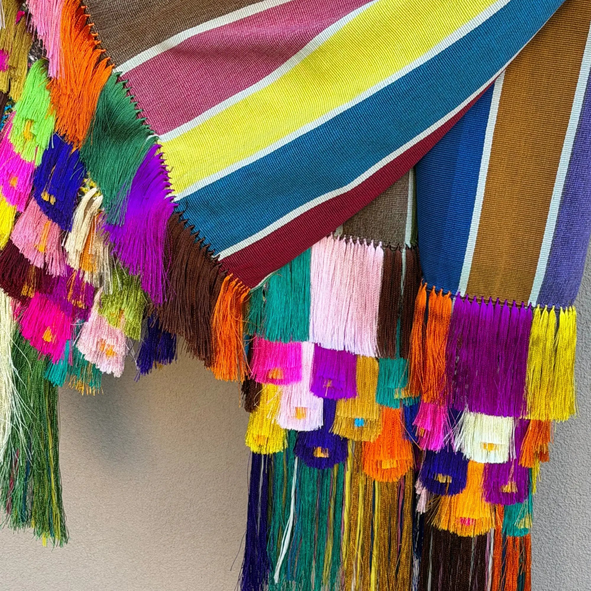 Vibrant, handwoven textile with bold horizontal stripes in pink, yellow, blue, and brown, adorned with cascading multicolored tassels.