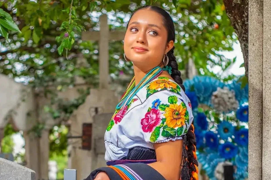 Woman wearing a traditional embroidered blouse with colorful floral patterns and a dark blue skirt adorned with gold lace trim.